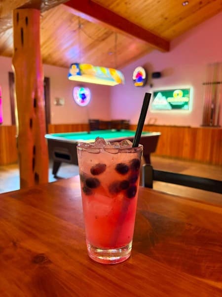 A tall berry cocktail on the bar rail with the pool tables and neon signs behind it