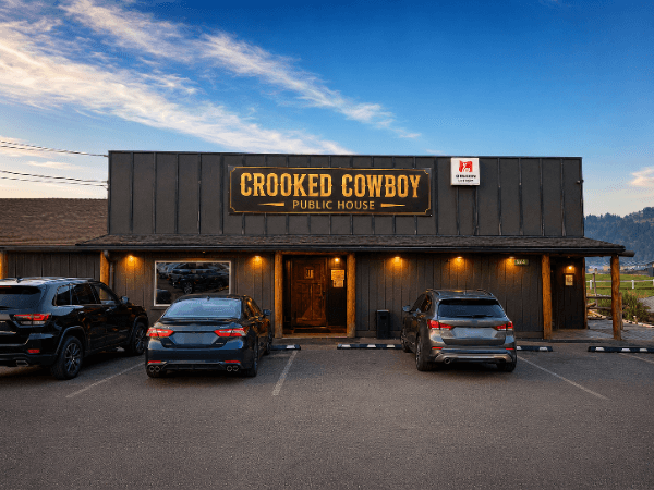 The Crooked Cowboy Public House at dusk — a dark timber storefront with the lit sign over the door and cars parked out front