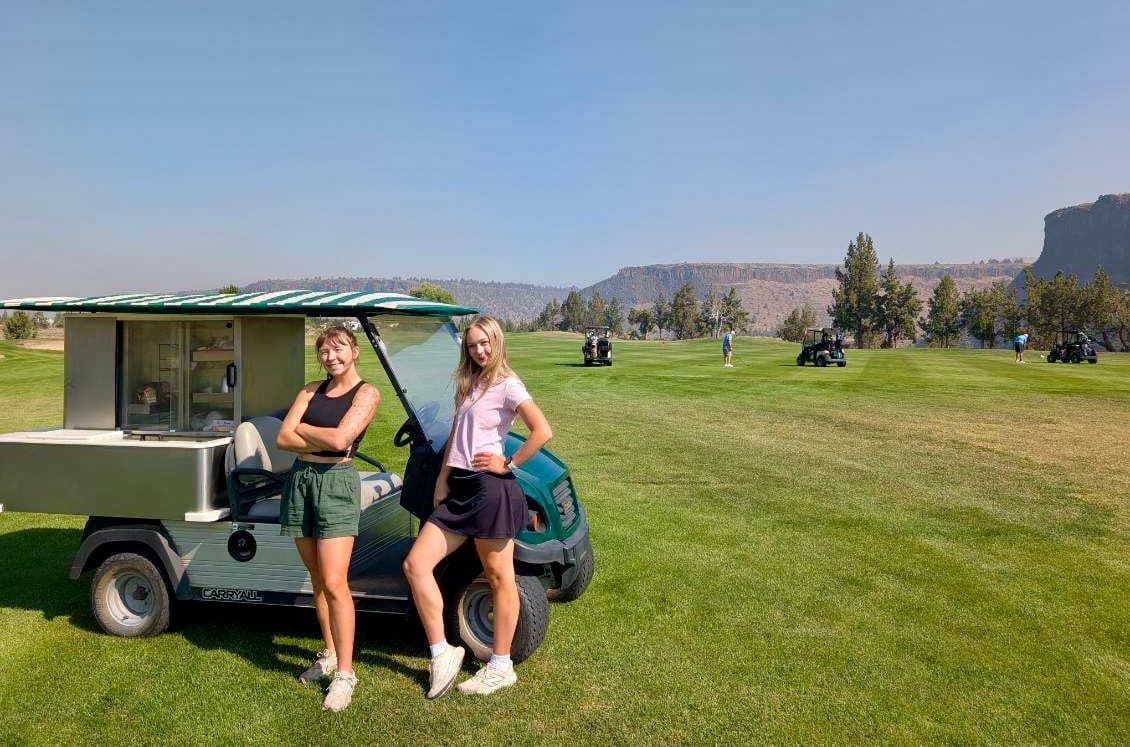 Two members of the Crooked Cowboy beverage cart crew standing beside the stocked cart on the fairway, with the canyon rim behind them